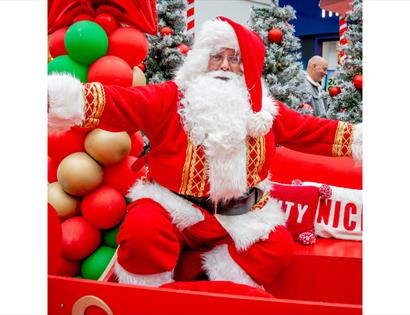 A person dressed as Santa Claus sits on a bright red sleigh surrounded by festive decorations. They wear a traditional red suit with white fur trim, g