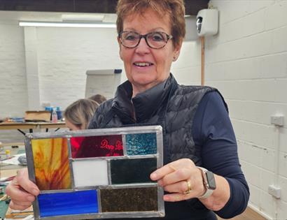 Person holding a handmade stained glass panel in a workshop filled with tools and materials.