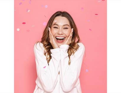 A young woman stands against a bright pink background, smiling widely with her eyes slightly closed and hands pressed to her cheeks in excitement. She
