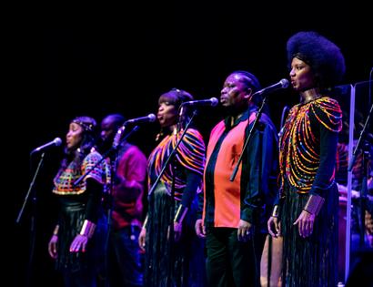 Performers sing into microphones on stage at De La Warr Pavilion, wearing vibrant costumes for Bob Marley reimagined concert.