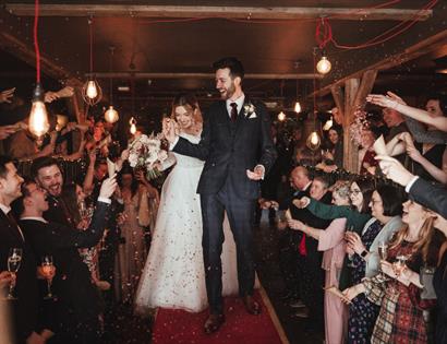 Couple walk through cheering wedding guests at a rustic venue, promoting The Bell’s Quirky Wedding Fair.