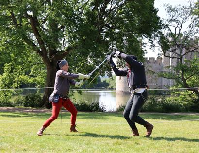 Call to Arms demonstrators stage medieval sword fighting on grass at Bodiam Castle, with the moat and stone walls behind.