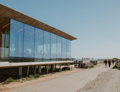 Modern glass-fronted Rye Harbour Discovery Centre beside coastal path, with visitors, seating area and sea views.