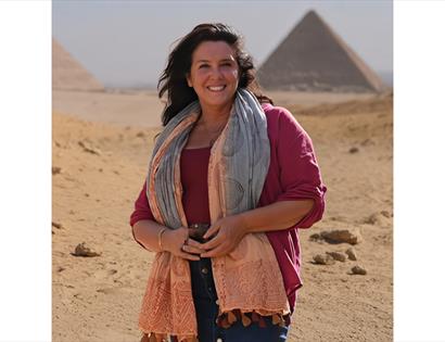 Portrait of Bettany Hughes in desert landscape with pyramids behind, promoting DLWP event.