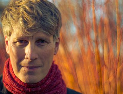 Close-up portrait of Scott Matthews outdoors wearing a red scarf, with short light hair, against a softly blurred background of warm-toned trees.