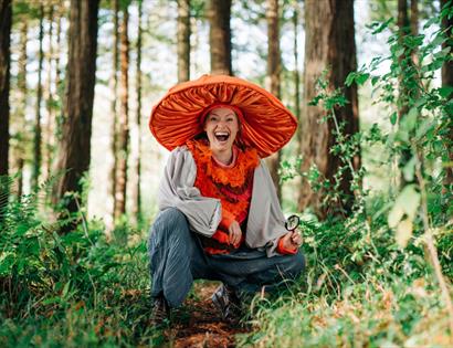Performer in oversized orange mushroom costume crouching in forest at Bedgebury, holding magnifying glass among trees.
