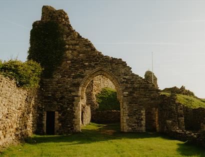 Stone archway and ivy-clad ruins of Hastings Castle in warm sunlight, with grassy grounds and hillside beyond.