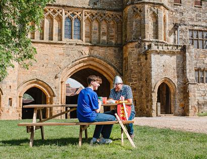 Two visitors sit at a picnic table beside the historic stone arches and medieval architecture of Battle Abbey.