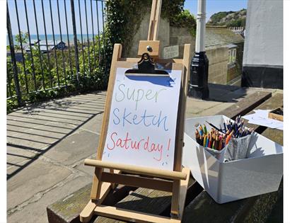 Easel sign reading “Super Sketch Saturday” beside art materials, overlooking the sea at Hastings Contemporary