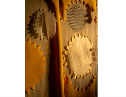 Abstract paper installation with layered cream, yellow and patterned cut-out shapes, lit by warm golden light.