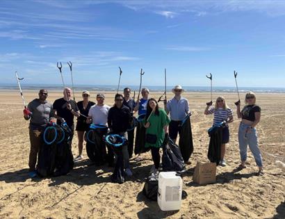 Community volunteers on a sandy beach holding litter pickers and rubbish bags during a community beach clean event.