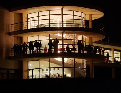 De La Warr Pavilion at night, glowing with interior lights and silhouetted visitors on curved balconies.