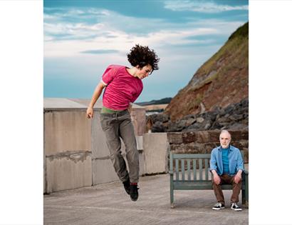 Outdoor dance duet at De La Warr Pavilion, with one performer leaping mid-air beside another seated on a bench by the sea.