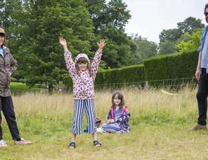 Family outdoors at Bateman's on a grassy field, with two adults and two children spaced apart, trees in the background on a cloudy day.