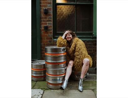 Person in a shaggy coat seated beside metal beer kegs in an urban alleyway with brick walls and green doors.