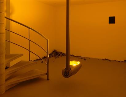 Warmly lit contemporary gallery installation featuring a spiral staircase, suspended light sculpture and scattered leaves.