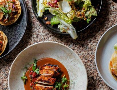 Overhead view of colourful dishes at Crafted, including tacos, grilled chicken, salad and golden empanadas.