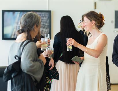 Guests with drinks chat at summer exhibition opening, De La Warr Pavilion, Bexhill, artwork in background.