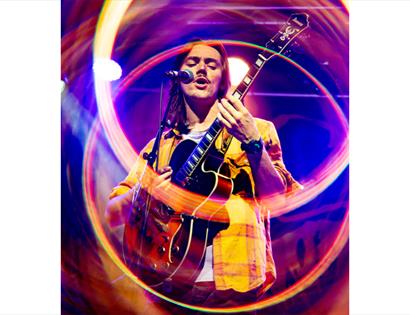 Performer sings while playing electric guitar on stage, surrounded by colourful light rings at Talent Accelerator Summer Sessions, DLWP.