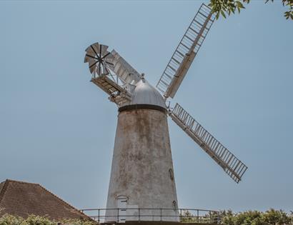 Stone Cross Windmill viewed from below, framed by leafy branches against a blue sky, with sails extended above a grassy area.