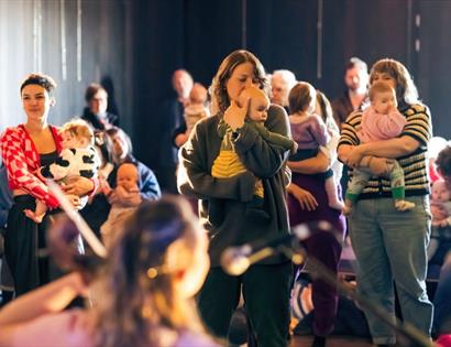 Caregivers holding babies gather around a live performance during A Dance for Us, a free event for babies 0-12 months.