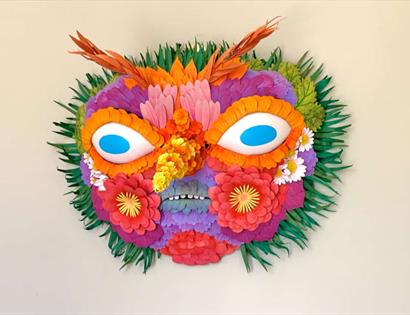 Bright floral mask made from layered paper flowers and leaves, with blue eyes, orange brows, and green leaf edges.