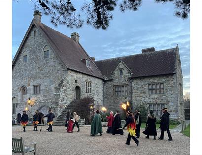 People in Tudor-style costume carrying flaming torches outside a historic manor house during a traditional wassail celebration.