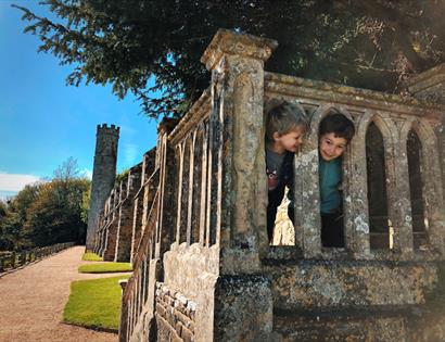 Children peeking through the stone balustrade at Battle Abbey, with historic walls and a tower along a sunny pathway.