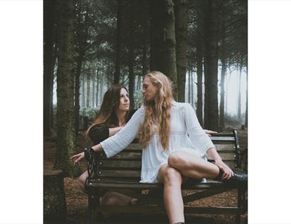 Promotional image for True Foxes at De La Warr Pavilion, showing two performers seated on a bench in a wooded setting.