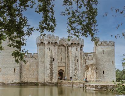 Bodiam Castle reflected in the surrounding moat, framed by tree branches under a bright sky.