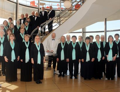 Bexhill Choral Society posed on a staircase at De La Warr Pavilion in concert dress, promoting a performance of Mendelssohn’s Elijah.