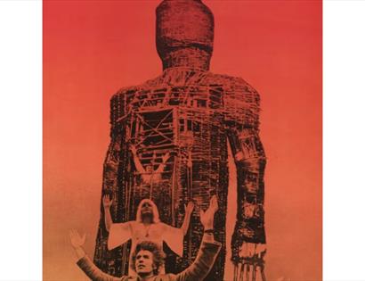 Promotional image for The Wicker Man showing a giant wicker figure towering above raised figures against a red sky.