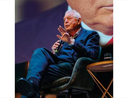 Speaker (John Simpson) on stage in armchair holding microphone , with large screen backdrop and table beside them.