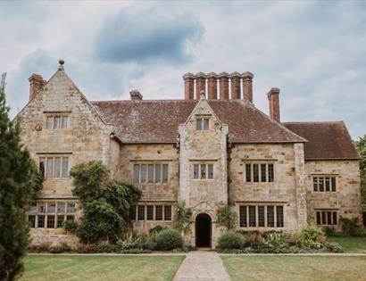 Batemans, a historic stone manor with tall chimneys and gardens, seen from the front on a cloudy day.