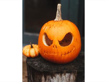 Halloween at The Hub, Bodiam: carved pumpkin with spooky face displayed on a rustic wooden stump.