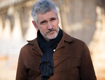 Portrait photo of a Curtis Stigers in a brown jacket and black scarf, standing outdoors with soft-focus background.