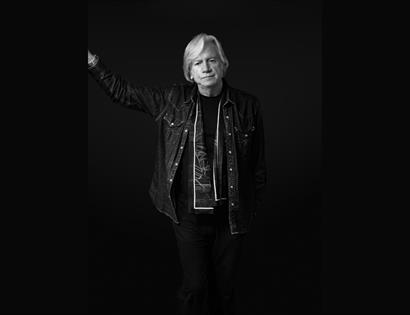 Black-and-white portrait for “The Voice of the Moody Blues: Justin Hayward,” musician with raised arm against dark background.