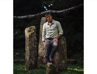 Seth Lakeman in a rolled-sleeve shirt leaning on ancient standing stones in a wooded outdoor setting.