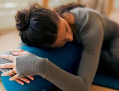 Person resting on a blue bolster while lying on a yoga mat in a relaxed pose indoors.
