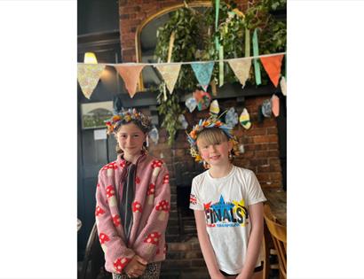 Two children wearing handmade flower crowns at a workshop in The Crown pub in Hastings, with bunting and decorations.