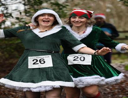 Festive holiday run: two women dressed as Santa jogging together on a forest path.