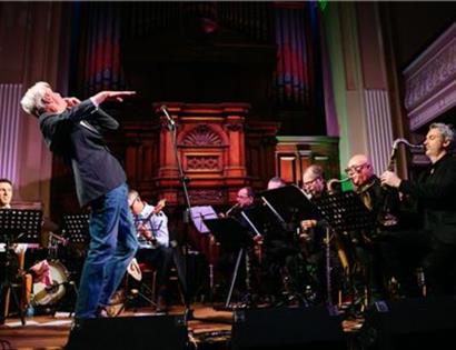 Musician performing energetically on stage with a live band in a grand hall featuring ornate wooden panels and organ pipes.