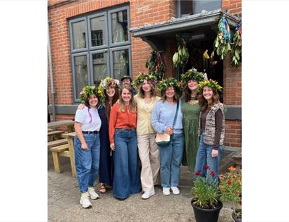 Group wearing colorful flower crowns outside The Crown in Hastings during a workshop, with plants and decorations nearby.