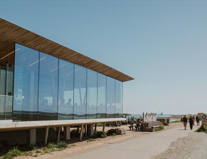 Modern glass visitor centre beside a coastal path at Rye Harbour Nature Reserve, with walkers, benches, and sea views.