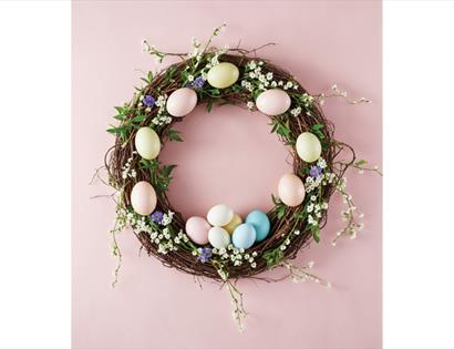 A spring wreath decorated with pastel eggs, greenery, and small flowers on a soft pink background.