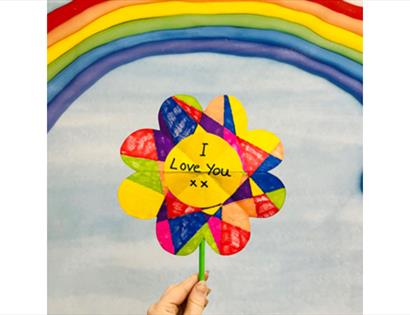 Hand holding a colourful heart-shaped pinwheel reading “I Love You xx” against a rainbow and soft blue sky background.