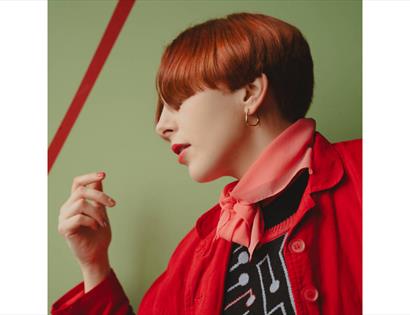 Image of Laura Fell with short auburn hair wearing a red jacket, pink scarf and patterned top against a green wall.