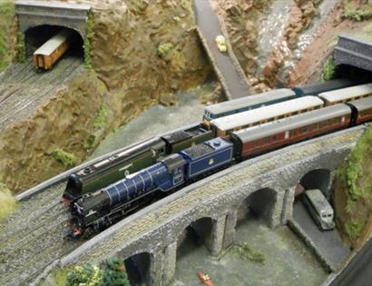 Model railway scene featuring a detailed steam train crossing a stone viaduct between two tunnels in a rocky landscape.