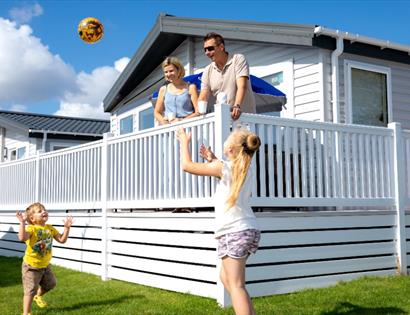 Image shows a caravan at Pevensey with 2 adults on the decking and 2 children playing a ball game on the grass