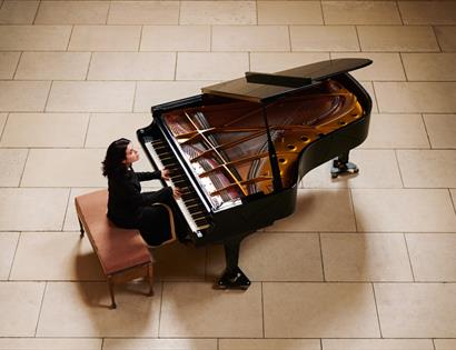 Person seated at a grand piano in a large tiled hall, playing with the piano lid open.